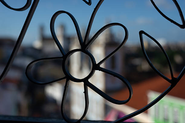 Lantern sits on a windowsill, casting a soft indoor contrast against backdrop of vibrant cityscape. Beyond glass, tiled rooftops and church rise into view, blending architectural charm of the city