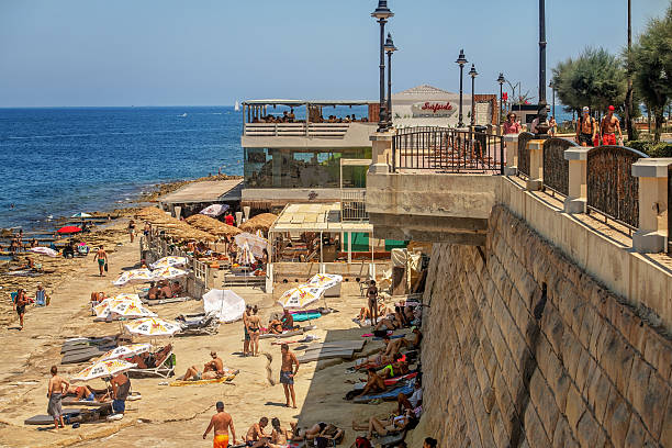 Sliema, Malta - 07.06.2025: Tourists sunbathe and swim at a rocky urban beach with sunbeds and straw umbrellas near a seaside café on a sunny summer day.