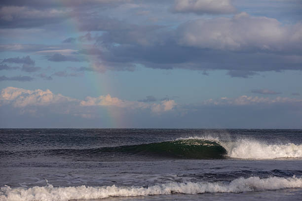A distant rainbow touches the horizon as a small clean ocean wave peels along the shore. Captured in magical afternoon light.