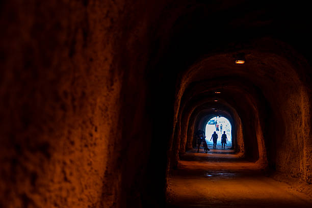 Dark rustic tunnel with rough earthy walls with several silhouetted figures walking towards the bright light at the far end. Montenegro, Budva, September, 20, 2024