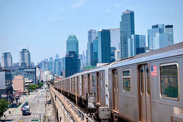 Queens, New York City, New York USA – July 7, 2025: A silver subway train glides westward above Queens Boulevard as a full Manhattan skyline unfolds behind it in the haze of midday light.