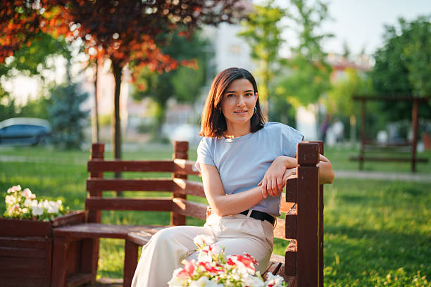 A young woman sits comfortably on a park bench and smiles warmly at the camera. The scene conveys a relaxed, friendly atmosphere in a peaceful outdoor setting.