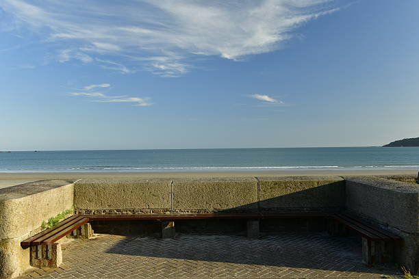A peaceful coastal setting featuring a pale green bench and table on grassy dunes facing the calm sea. Bright blue sky, distant horizon, and windswept coastal vegetation create a serene, outdoorsy mood perfect for travel, relaxation, and nature keywords in Denmark.