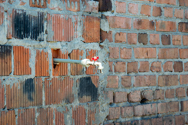 A water spigot is installed through a hole in a brick wall at a construction site under bright daylight.