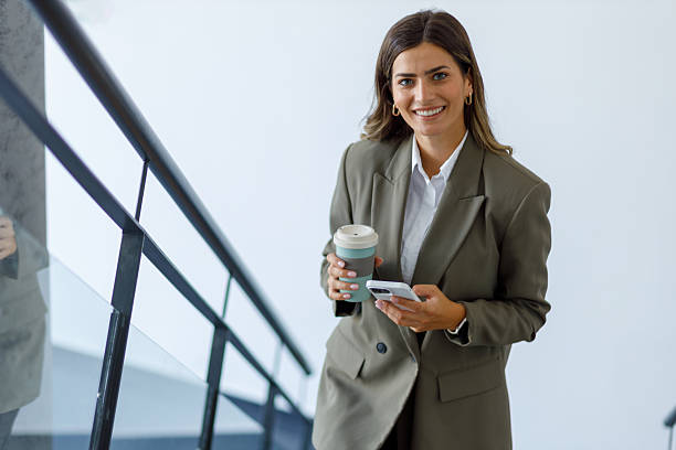 istock Smiling businesswoman walking in office hallway, holding reusable coffee cup and using smartphone 2224010477