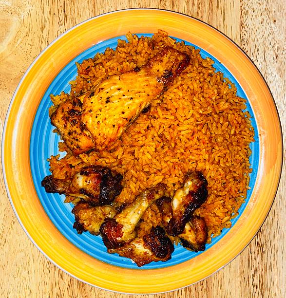 A colourful plate of food on a wooden table, featuring seasoned chicken pieces (including a drumstick and wings) served over a bed of vibrant Jollof rice, seasoned with spices.