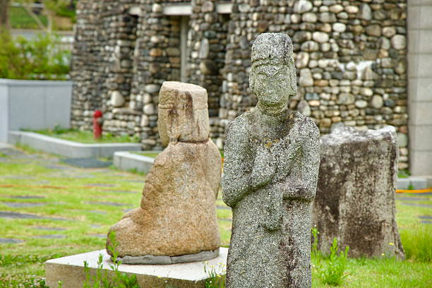 Buyeo County, South Korea - June 13th 2025: A closeup view of a weathered stone statue with folded arms displayed outside Buyeo National Museum, with a rock wall and lawn in the background.