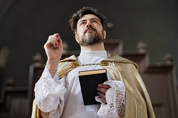 Caucasian middle aged man wearing priest vestments standing in church holding book and raising hand while looking upward, facial hair visible, wooden background
