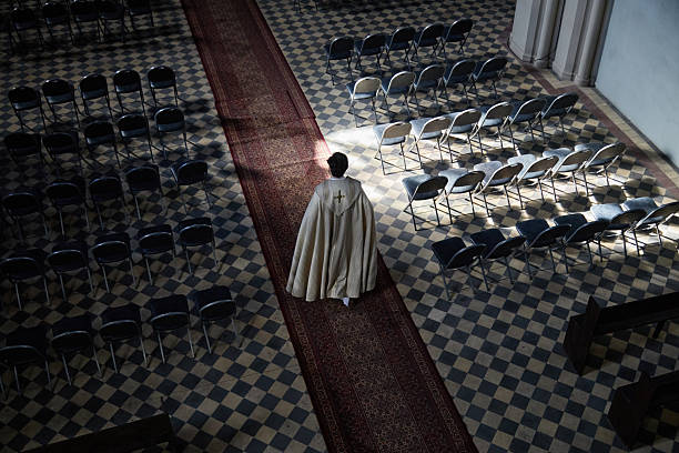 Caucasian middle aged male priest wearing robe walking down church aisle between rows of empty chairs, back view, sunlight streaming onto patterned floor, religious setting