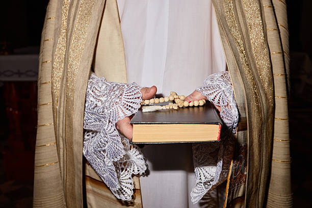 Man holding prayer beads and book while standing in church, wearing ornate religious vestments, hands visible with lace sleeves, participating in religious ceremony