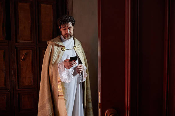 Middle aged Caucasian man wearing priest robe standing in church holding smartphone, looking at device with serious expression, wooden doors and religious setting in background