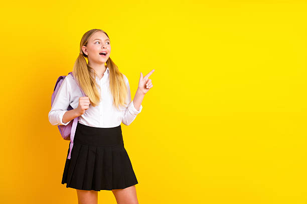 istock Smiling student in a white shirt holding a backpack and pointing with excitement against a vibrant yellow background 2224050419