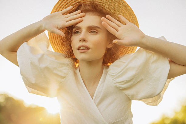 A young woman with curly hair stands gracefully in a sunlit field, wearing a flowing white dress and a straw hat. The golden hour light casts a warm, ethereal glow, highlighting her serene expression and the natural beauty of the scene. Her relaxed pose and the surrounding landscape evoke a sense of peace and timeless elegance.