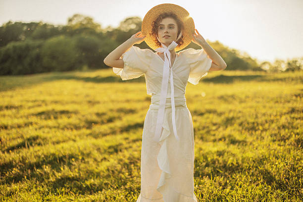 This evocative image depicts a young woman with curly red hair walking through a sunlit meadow during late afternoon. Her vintage-style white dress, with puffed sleeves and a tie at the neck, adds a timeless elegance to the scene. She appears absorbed in the moment, gently touching the wildflowers as she walks by. The warm, natural lighting and the open field background enhance the pastoral and peaceful ambiance, evoking a sense of freedom and connection to nature.