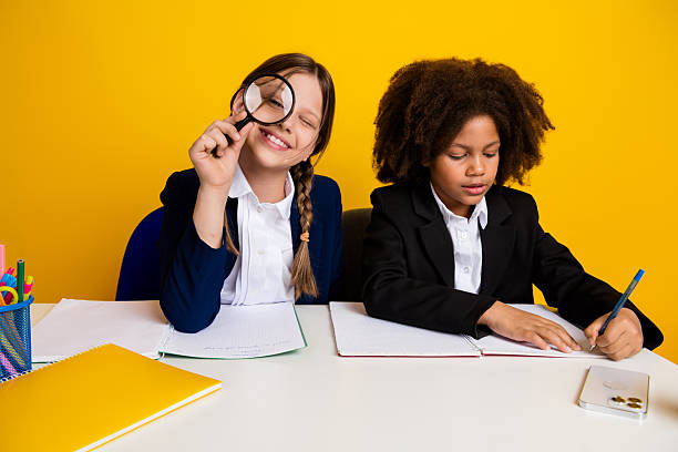 istock Two cheerful schoolgirls engaging in fun learning at a desk, featuring a bright yellow background and youthful interaction 2224054025