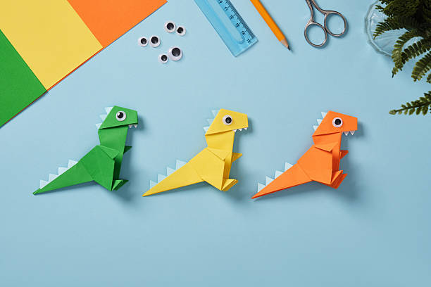 istock Origami work table with sheets of paper, googly eyes, scissors, ruler, and eraser on a light blue background. Finished paper dinosaur toys. 2224056906