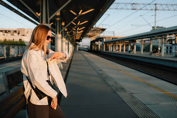 istock Stylish woman checking time on her watch while waiting for train at sunset, standing on platform of railway station with train arriving in background. Woman glancing at watch, waiting for train 2224067383