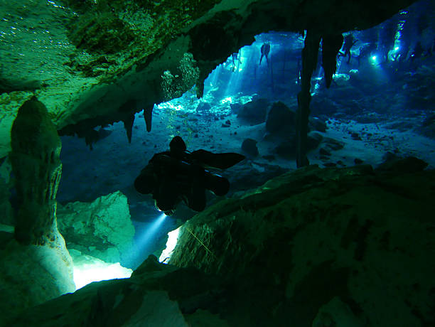 A scuba diver explores an underwater cenote cave with stalactites and blue light filtering through the water, Riviera Maya, Mexico