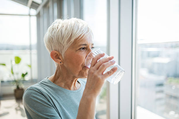 istock Senior woman drinking water in a bright, indoor setting by a window 2224075515