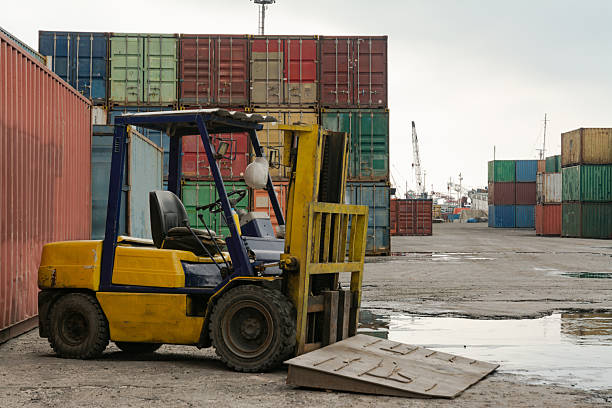 A high-resolution image of neatly stacked shipping containers at a container port or shipyard captured during midday under a cloudy sky. A parked forklift is visible near the containers, adding a human element and emphasizing the scale and functionality of the logistics environment. The overcast lighting softens shadows and highlights the industrial textures, creating a balanced and professional look. Ideal for illustrating global trade, cargo handling, and port operations. Perfect for use in editorial content, logistics marketing, or supply chain visuals that showcase real-world freight infrastructure.