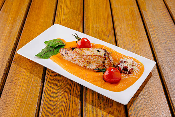 A beautifully plated grilled fish fillet rests on a vibrant squash puree, accompanied by roasted cherry tomatoes and fresh basil. The dish showcases culinary artistry on a wooden table.