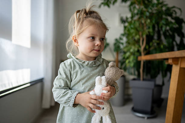 Adorable toddler girl standing in a modern living room, holding her beloved teddy bears, enjoying a quiet moment of play and affection in a cozy domestic setting