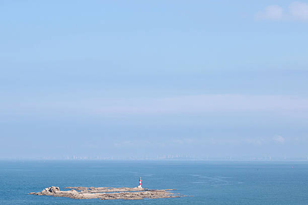 This image showcases a tranquil coastal scene with a vibrant lighthouse stationed on a rocky island amidst expansive ocean waters under a clear blue sky.