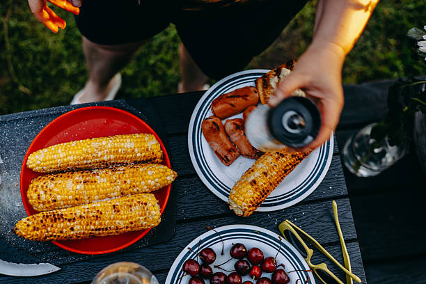 Delicious barbecue spread with grilled corn, sausages, cherries, ready for a summer gathering. A person seasons grilled corn on the cob on a black outdoor table, ideal for a summer barbecue party.