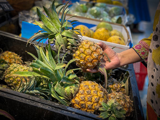 A close-up view captures a human hand reaching into a display bin to carefully select a ripe, yellow pineapple at a modern supermarket or grocery store. Several other fresh pineapples are visible, emphasising healthy eating, the choice of natural and tropical fruits, and the everyday experience of grocery shopping for quality produce