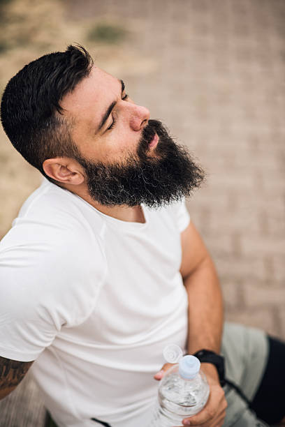 A young man sits on a stadium bench, water bottle in hand, seen from above. With his eyes closed and a calm expression, he takes a quiet break after running, enjoying the warmth of the sun and a moment of stillness.