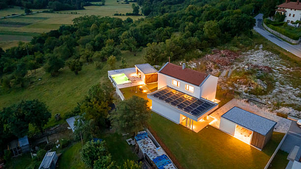 Aerial photograph of an energy-efficient modern residence featuring rooftop solar panels, warm interior illumination, and minimalist white exterior nestled in rolling green countryside at dusk.