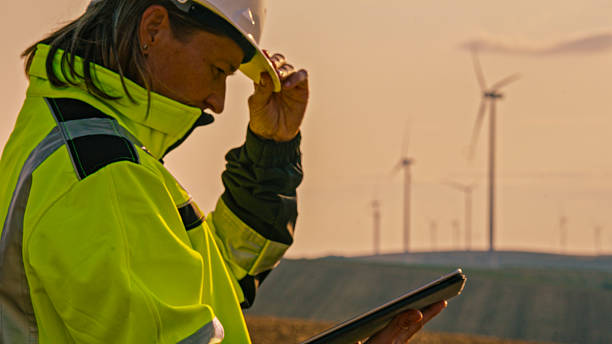 istock Female Engineer Inspecting Wind Turbines at Sunset Holding Digital Tablet in Bright Yellow Jacket 2224108216