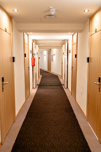 A hotel corridor with a symmetrical design featuring closed wooden doors lining both sides. Soft ambient lighting illuminates the scene, creating a cozy and inviting atmosphere. The hallway is carpeted with a visible fire extinguisher mounted on the wall, adding a practical element to the decor.