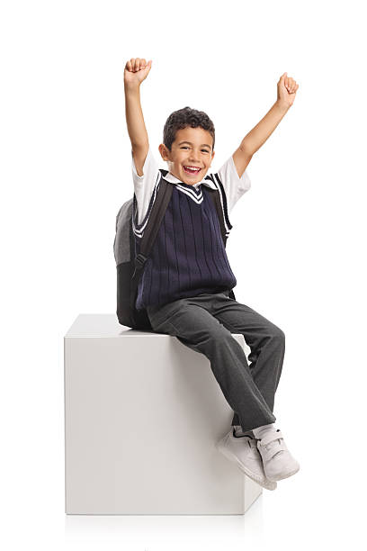 Smart elementary age schoolboy sitting at a desk in a modern classroom, raising his hand enthusiastically to answer a teacher's question during an engaging lesson