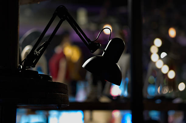 A dark, silhouetted desk lamp with a classic bell-shaped shade stands in stark contrast to a distant, glowing circular light. The blurred background enhances the minimalist composition, drawing attention to form, depth, and quiet atmosphere.