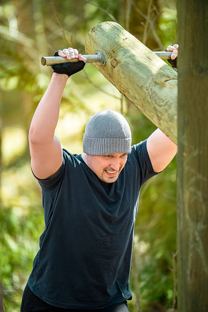Man lifting heavy wooden log outdoors during functional workout. Dressed in a beanie and gloves, showing strength and determination. Fitness and outdoor training.