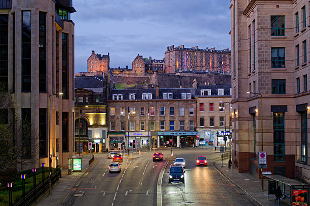 Cars with lights at T junction in Edinburgh at dusk. View towards illuminated historic Edinburgh castle and modern office buildings. Edinburgh, UK, 05.04.2025