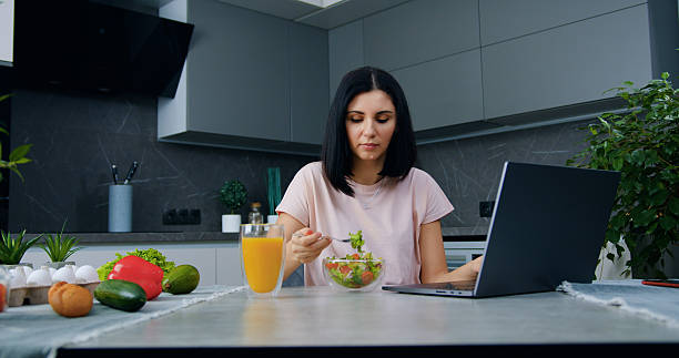 istock Attractive concentrated 30-aged black-haired woman sitting at kitchen table ,reading information on laptop and eating vegetable salad 2224149839