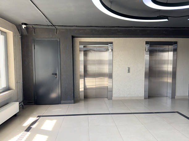 istock Modern building interior with two stainless steel elevators, dark utility door, surveillance camera and geometric ceiling light, illustrating clean minimalist hallway in residential or office space 2224154672
