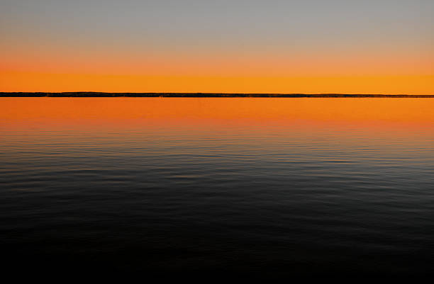 Serene view of a calm lake reflecting a vibrant sunset. The sky features a beautiful gradient from deep blue to warm orange, creating a peaceful and minimalist landscape.