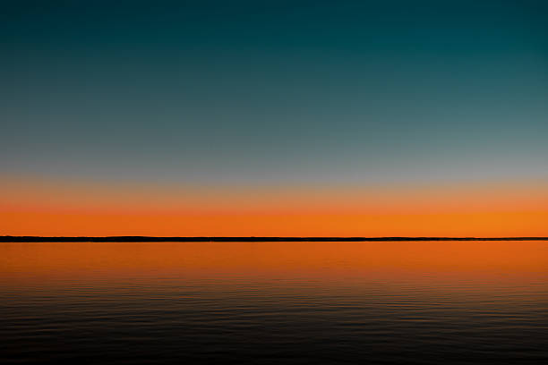 Serene view of a calm lake reflecting a vibrant sunset. The sky features a beautiful gradient from deep blue to warm orange, creating a peaceful and minimalist landscape.