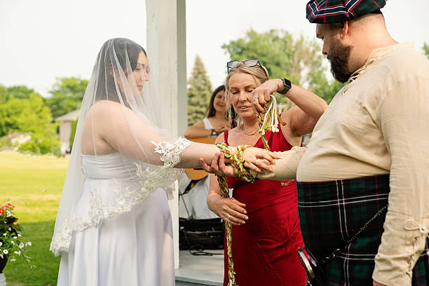 Millennial wedding ceremony outdoors in summer. One spouse is a non-binary person, dressed in a Scottish kilt with a beret, body positive bride dressed in white, celebrant is wapping the arms of the spouses with a rope according to ancient Celtic tradition. Horizontal waist up outdoors shot. This was taken in Quebec, Canada.