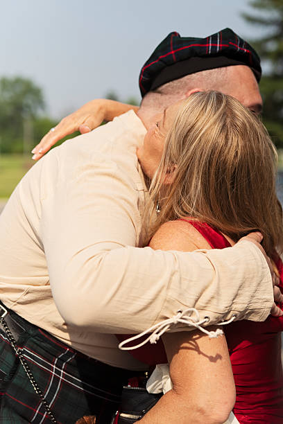 Millennial non-binary person greeting mother before wedding. They are dressed in a Scottish kilt with a beret, body positive with short hair and a beard. Mother is wearing a red dress. Vertical waist up outdoors shot. This was taken in Quebec, Canada.