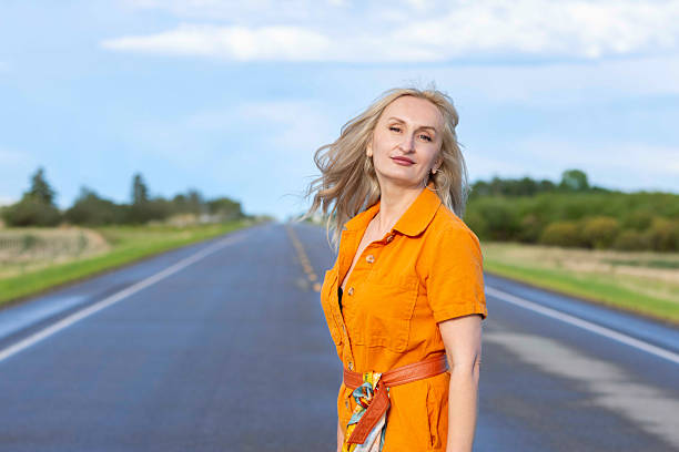 Young blond woman with long hair in retro orange jumpsuit is on the country empty highway traveling in summer.