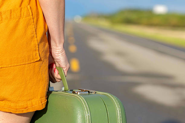 Woman in orange jumpsuit on a highway, hand is holding a vintage old green suitcase. Hitchhiker on a rural empty road.
