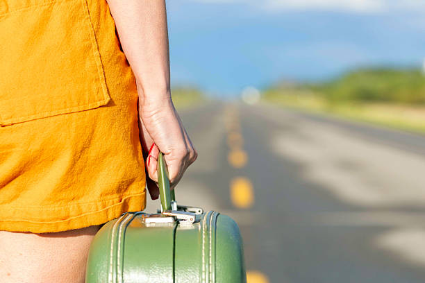 Woman in orange jumpsuit on a highway, hand is holding a vintage old green suitcase. Hitchhiker on a rural empty road.