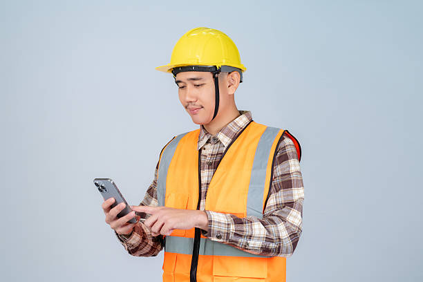 istock Smiling young Asian engineer or worker wearing yellow hard hat and reflective vest using smartphone working on grey background. 2224177342