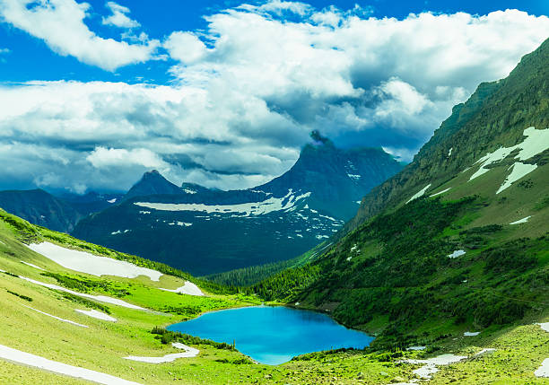 A vivid view of Ptarmigan Lake glowing with surreal turquoise blue beneath the towering peaks of Glacier National Park, Montana. The contrast of lingering summer snow, vibrant alpine meadows, and dramatic clouds creates a scene that captures the untamed beauty of the northern Rockies.