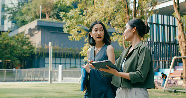 istock Two young Asian businesswomen discussing work while holds digital tablet and carries documents while walking outside office building. Sustainable business outdoor. 2224192081