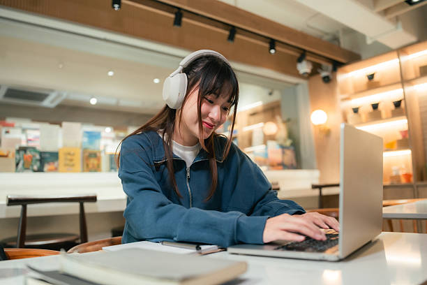 Gen Z woman multitasks in a co-working space, listening to an audiobook, taking notes in her book, and studying with her laptop